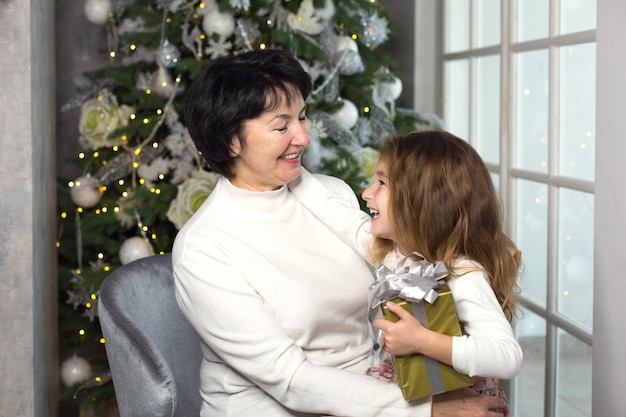 Abuela con una niña en el fondo de adornos navideños y un gran ventanal.