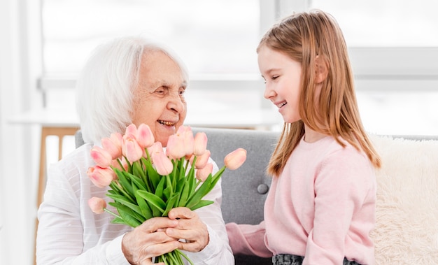 Abuela con ramo de flores de tulipán en sus manos sentada con su nieta en el sofá y sonriendo