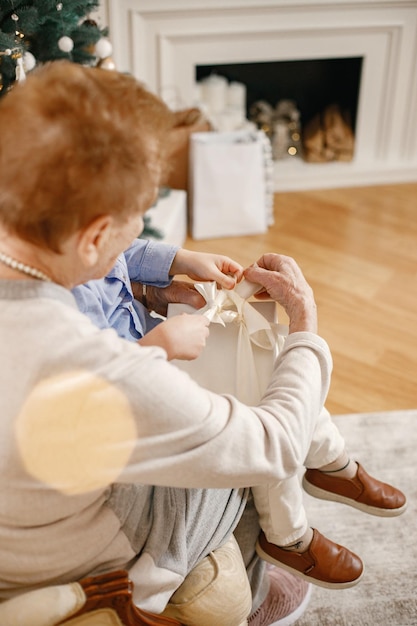 Abuela con su nieto el día de Navidad. Anciana y niño abriendo un regalo. Mujer y niño con ropa beige y azul.