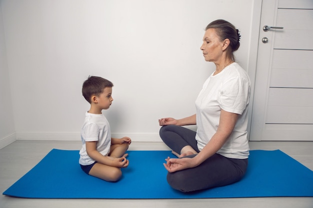 La abuela y su nieto están sentados en una estera de yoga en un apartamento blanco en casa y meditando en la posición de loto