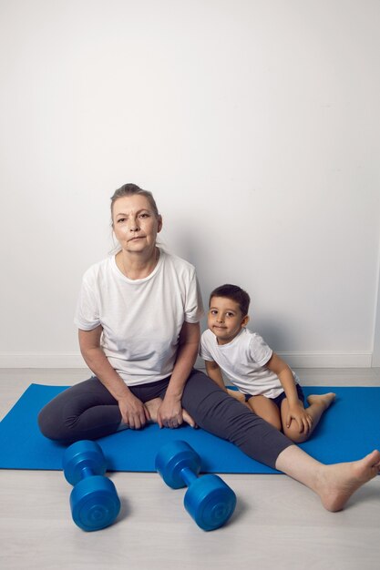 La abuela y su nieto están sentados en una estera de yoga en un apartamento blanco