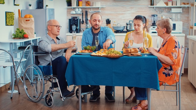 Abuelo con discapacidad en silla de ruedas y familia cenando. Dos parejas felices hablando y comiendo durante una comida gourmet, disfrutando del tiempo en casa sentados alrededor de la mesa de la cocina.