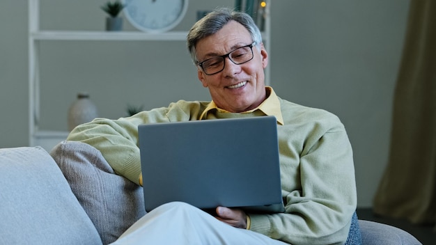 Abuelo feliz sentarse en el sofá de la sala de estar hombre caucásico anciano maduro mirando el trabajo de la computadora portátil de pantalla