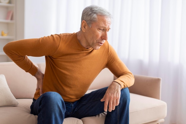 El abuelo infeliz sentado en el sofá y frotándose la parte inferior de la espalda