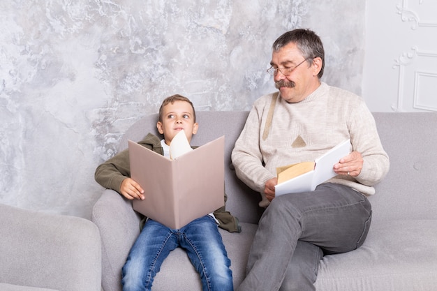 Abuelo y nieto están sentados en la sala leyendo libros juntos. Boy y su sonriente abuelo pasan tiempo juntos en el interior. Senior hombre con un niño