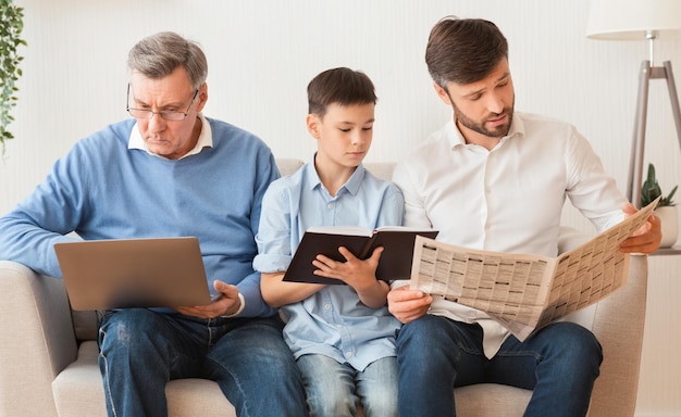 Abuelo, padre e hijo usando laptop, leyendo libros y periódicos sentados en el sofá en casa. Actividades de fin de semana de la familia masculina.