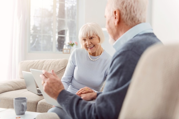 Abuelos cariñosos. Agradable anciano mostrando a su esposa la tableta con las fotos de su nieto mientras la mujer la mira con una mirada divertida