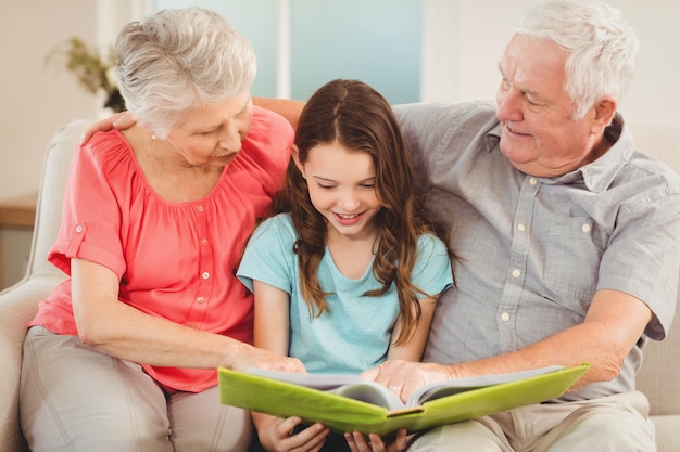 Abuelos y nieta sentados en el sofá y leyendo un libro con nieta