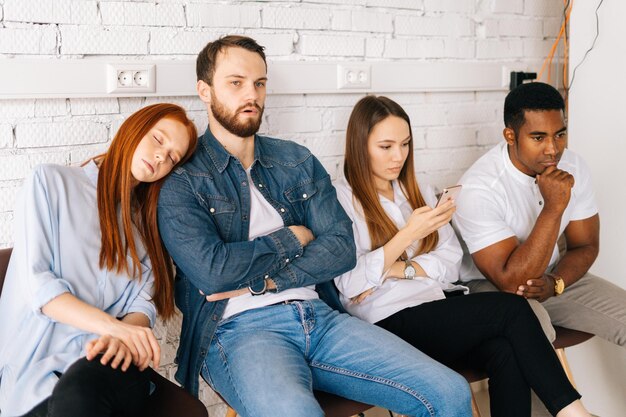 Aburridos, cansados, jóvenes y diversos candidatos de trabajo multiétnicos con ropa informal esperando una entrevista con hr, sentados en fila de cola en sillas en el moderno vestíbulo de la oficina sobre el fondo de una pared de ladrillo blanco.