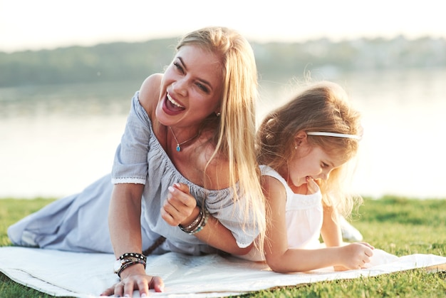 Acabei de ouvir uma piada. Rindo. Foto de uma jovem mãe e sua filha se divertindo na grama verde com um lago ao fundo