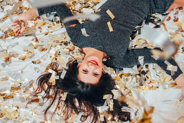 Se acabó la fiesta. Libre solo. Morena acostada en la cama salpicada de confeti. Mujer feliz y sonriente. Recuerdos y relajación.