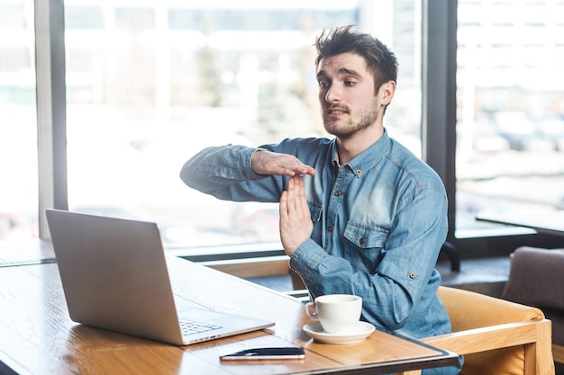 ¡Se acabó el tiempo! El retrato de vista lateral de un joven autónomo barbudo y apuesto con pantalones vaqueros azules está sentado en un café y haciendo una videollamada en una laptop con los brazos levantados y mostrando un gesto de pausa. Interior,