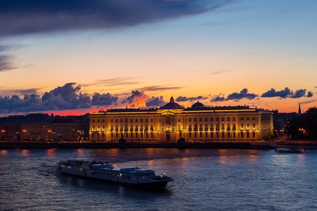 Academia de Artes de Rusia iluminada en San Petersburgo, Rusia. Barco a la deriva borrosa en medio del río. Hermoso paisaje al atardecer. Destino de viaje. Lugar de vacaciones. Concepto de pasión por los viajes.