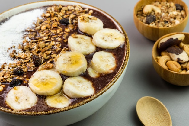 Açaí brasileiro em uma tigela branca com granola de mel de leite em pó de banana e nozes em pequenas tigelas de bambu Castanha de caju e fundo cinza do Pará Foco seletivo