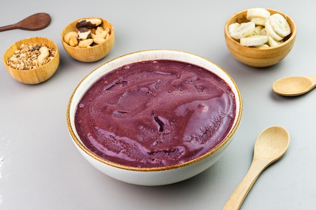 Açaí brasileiro em uma tigela branca granola de banana adicional e nozes em pequenas tigelas de bambu Caju e nozes do Pará Vista superior Fundo cinza Foco seletivo