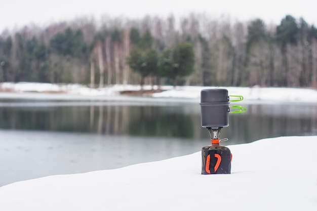 Acampamento de inverno ao ar livre contra o fundo da neve da floresta e do lago queimador de gás turístico com panelas para cozinhar