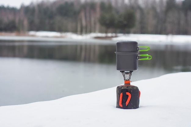 Acampamento de inverno ao ar livre contra o fundo da neve da floresta e do lago queimador de gás turístico com panelas para cozinhar