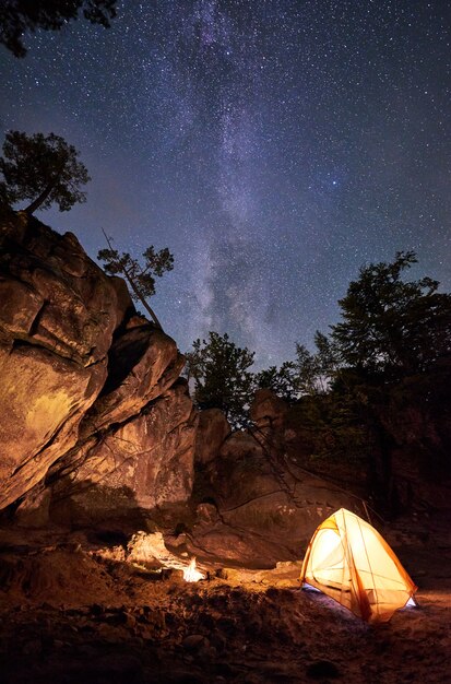 Acampamento de montanha à noite de verão em meio a uma enorme formação rochosa íngreme. pequena barraca do turista iluminada pela fogueira em chamas sob o céu estrelado escuro claro com a via láctea. conceito de turismo, caminhadas e viagens