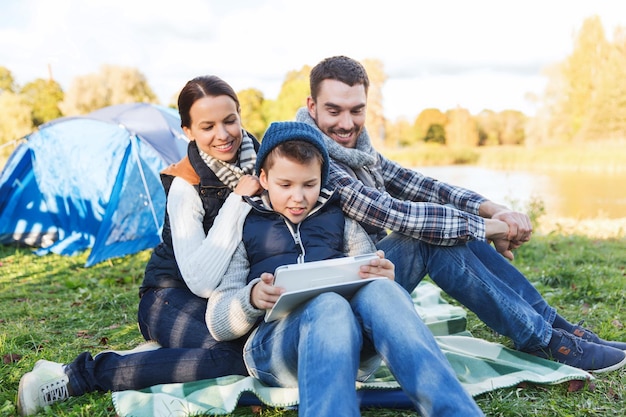 acampamento, turismo, caminhada, tecnologia e conceito de pessoas - família feliz com computador tablet pc e barraca no acampamento