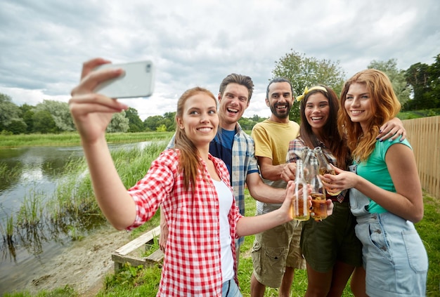 acampamento, viagens, turismo, caminhada e conceito de pessoas - amigos felizes com garrafas de vidro bebendo cidra ou cerveja e tomando selfie por smartphone no acampamento