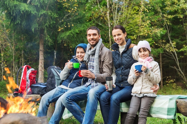 acampamento, viagens, turismo, caminhada e conceito de pessoas - família feliz sentada no banco e bebendo chá quente de xícaras na fogueira na floresta