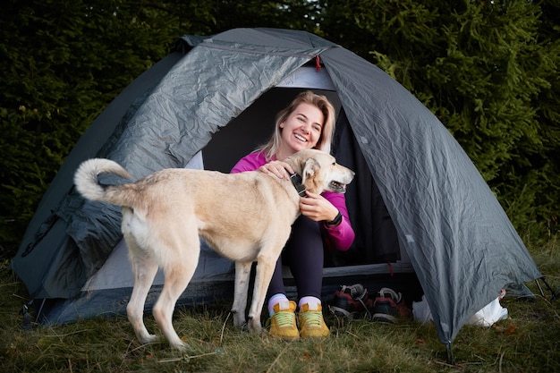 Acampar al aire libre Joven mujer feliz con una sonrisa en la cara sentada en una carpa turística gris y acariciando a su perro Vacaciones activas de excursionista con mascota en la naturaleza