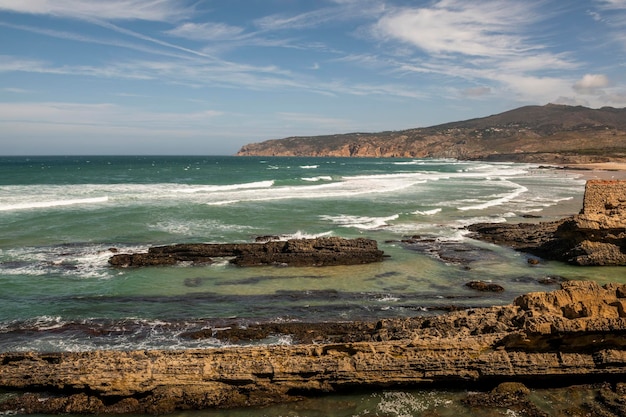 Acantilado de roca suelta y olas persistentes en la costa atlántica portuguesa y una playa con bañistas en un gran día de verano