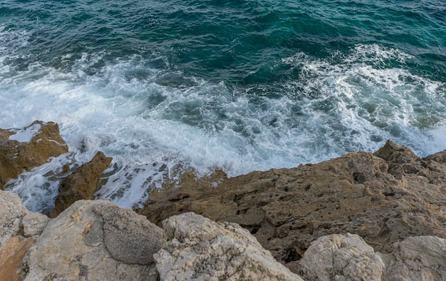 Acantilado de verano junto al Mar Mediterráneo, fuertes olas rompen con las rocas y dejan colores azules y turquesas junto con la espuma del mar.
