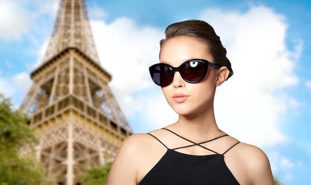 accesorios, anteojos, moda, gente y concepto de lujo - hermosa mujer joven con elegantes gafas de sol negras sobre el fondo de la torre eiffel de parís