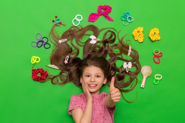 Accesorios para el cabello Una niña feliz yace rodeada de bandas elásticas y pinzas para el cabello y da un pulgar hacia arriba Peinados para niños Fondo verde aislado