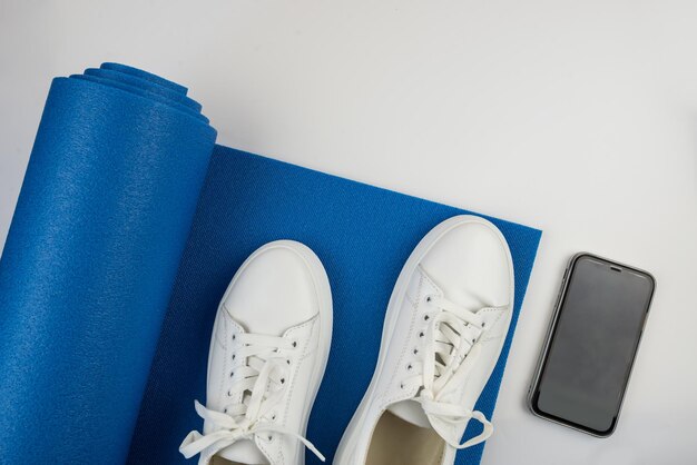 Accesorios de fitness concepto Foto de pesas teléfono inteligente con auriculares zapatillas de deporte blancas alfombra deportiva sobre un fondo blanco con espacio vacío