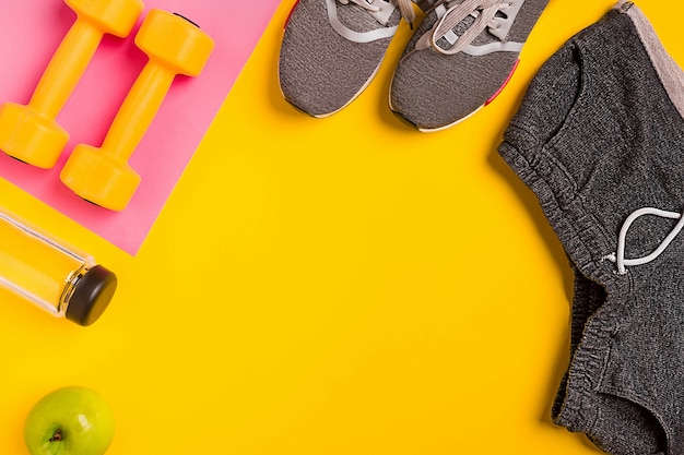 Accesorios de fitness sobre un fondo amarillo. Zapatillas, botella de agua, manzana y mancuernas. Naturaleza muerta