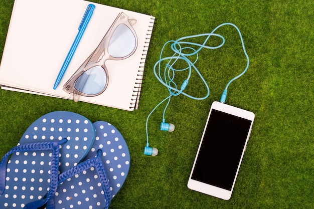 Accesorios de moda femenina, chanclas, teléfono inteligente con auriculares, bloc de notas, gafas de sol en la hierba