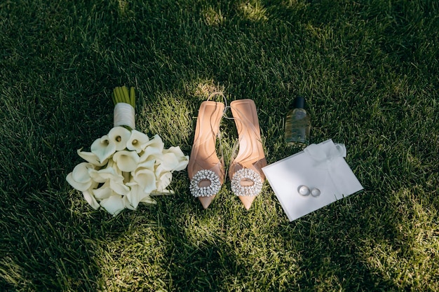 Accesorios de novia con ramo de lirios blancos calas zapatos y perfume en la hierba verde bajo el sol