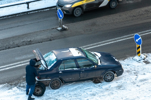 Accidente automovilístico en la carretera en invierno El conductor cambia la rueda en la carretera en la ciudad en el frío en condiciones difíciles Condiciones peligrosas de la carretera en condiciones heladas