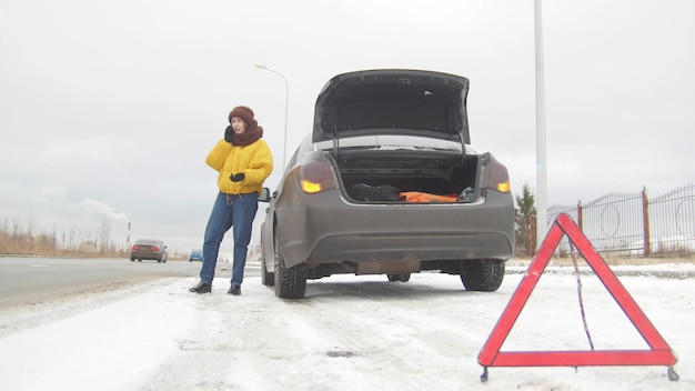 Un accidente automovilístico una mujer joven de pie junto a un coche averiado y pide ayuda por teléfono