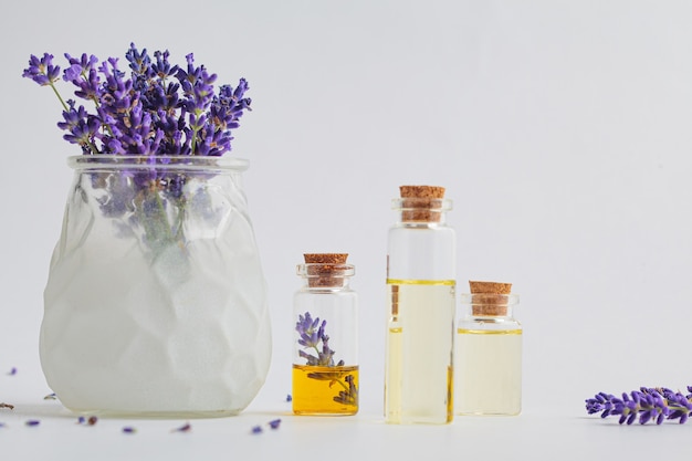 Aceite esencial de lavanda en pequeñas botellas de vidrio y flores de lavanda en un mortero, fondo blanco.