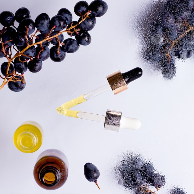 Aceites cosméticos de vidrio pipetas y botellas con uvas negras alrededor sobre fondo trasnlucid húmedo