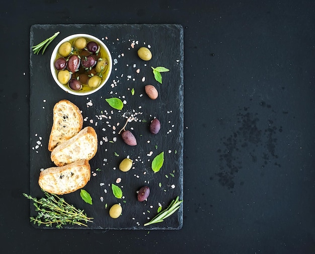 Aceitunas mediterráneas con hierbas y rodajas de ciabatta en bandeja de pizarra negra sobre la vista superior de fondo grunge oscuro