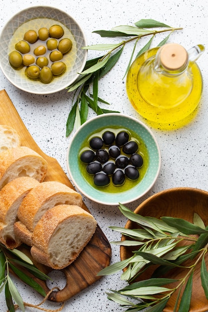 Aceitunas verdes y negras con aceite de oliva en una botella de vidrio, ramitas de olivo y pan ciabatta fresco cortado sobre tabla de cortar de madera. Fondo rústico blanco, concepto de comida mediterránea, vista superior