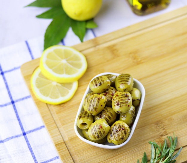Aceitunas verdes a la parrilla con romero, aceite de oliva y limón en un plato sobre tabla de cortar de madera en la mesa