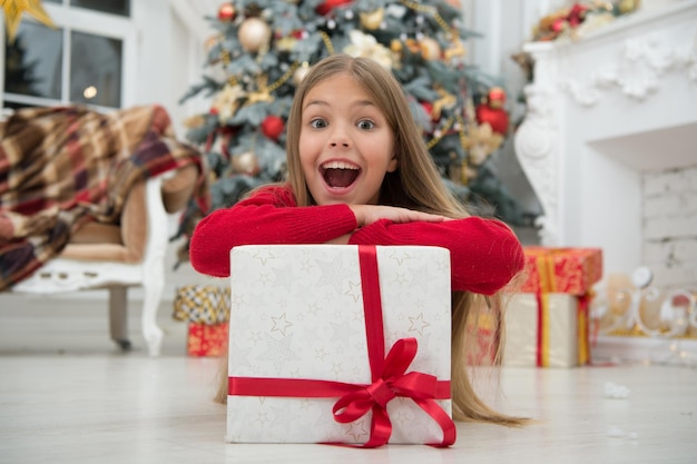 Se acerca la Navidad Pequeña niña linda recibió un regalo navideño Los mejores regalos de Navidad Niña pequeña con vestido elegante y caja de regalo Fondo de árbol de Navidad Niño emocionado por desempacar su regalo