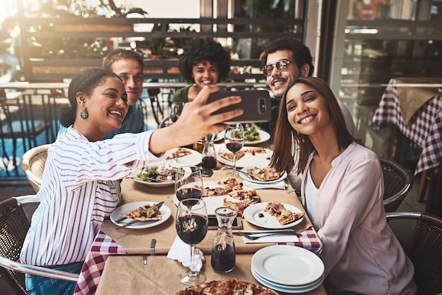 Acércate a la foto Captura de un grupo de alegres jóvenes amigos tomándose un autorretrato junto con un teléfono celular mientras están sentados en una mesa dentro de un restaurante
