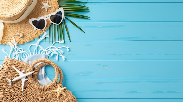 Acessórios de férias na praia, como chapéu de palha, óculos de sol, saco e folha de palma, dispostos em uma superfície de madeira azul vibrante, simbolizando viagens de verão e lazer