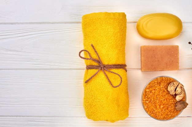 Acessórios de spa de banho plana leigos na mesa de madeira branca. Toalha amarela enrolada com sabonete e esponja.