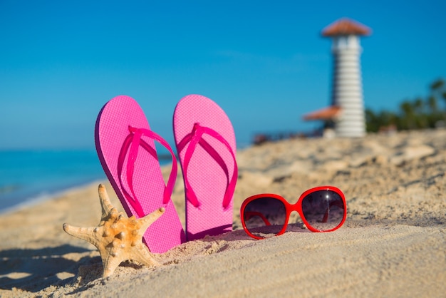 Acessórios marinhos femininos: sandálias, óculos de sol e estrelas do mar na praia de areia tropical contra o fundo do farol
