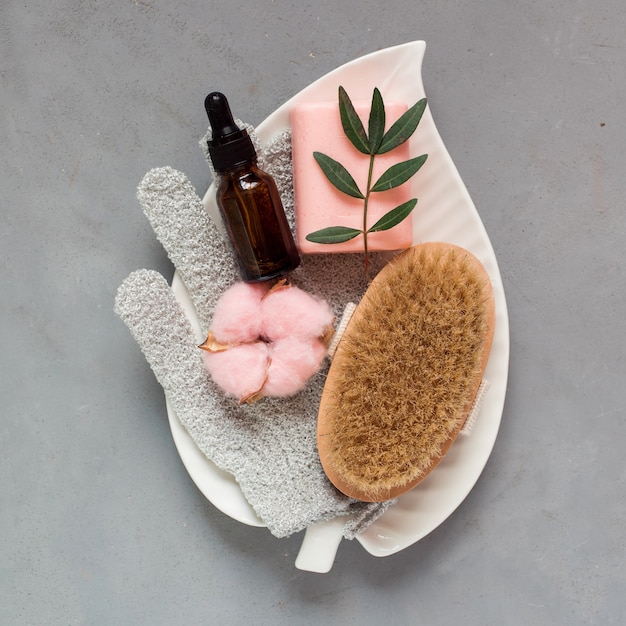 Acessórios para cuidados com a pele massageando escova em forma de luva de sabonete soro ou óleo no frasco na placa em forma de folha na vista superior do plano de fundo cinza