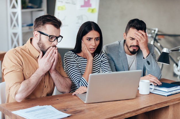 Ach nein! Drei frustrierte junge Geschäftsleute in eleganter Freizeitkleidung, die auf den Laptop schauen und Negativität ausdrücken