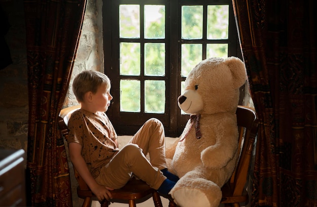 Achtjähriger trauriger Junge mit einem großen Teddybären nahe dem Fenster