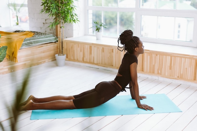 Achtsame afroamerikanische Frau mit dunkler Haut und engen Augen macht Yoga-Übungen im Innenbereich. Entspannen Sie sich und dehnen Sie sich in Kobra-Pose